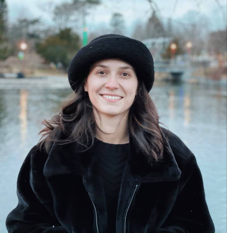 A photo of Mallory standing outside in front of a pond wearing a black hat, sweater, and jacket.