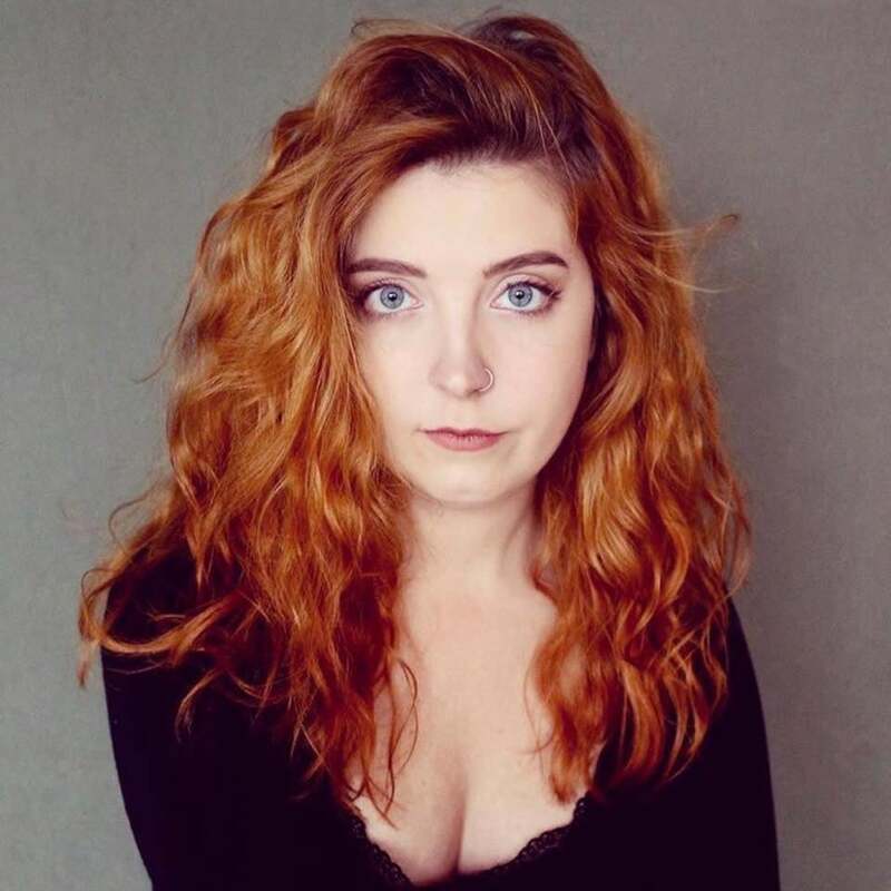 A headshot of Alison in front of a grey background. Alison has long red hair and is wearing all black.