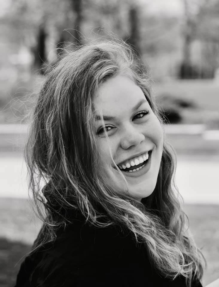 Black and white image of Erika Squires smiling in an outdoor setting. Erika has light curly hair, light eyes and dimples. Erika is wearing an oversized denim shirt and looks back over their right shoulder.